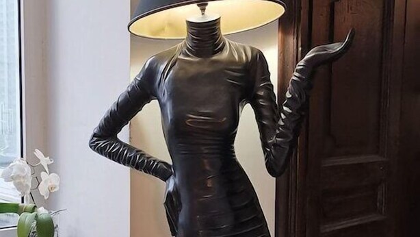 A sculpture of a human figure with a lamp on its head, placed in a corner of a room with a wooden door and a small table with a plant and a board game. - Costantinapoli 27 (Naples)