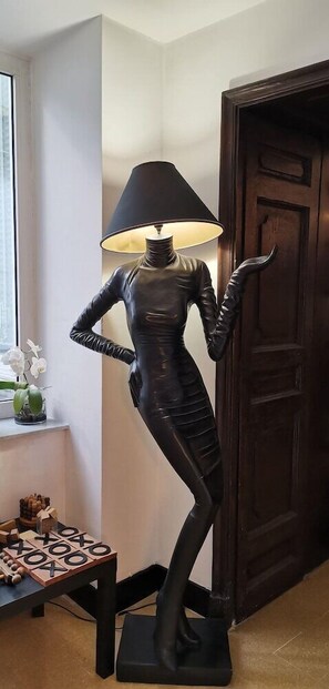 A sculpture of a human figure with a lamp on its head, placed in a corner of a room with a wooden door and a small table with a plant and a board game. - Costantinapoli 27 (Naples)