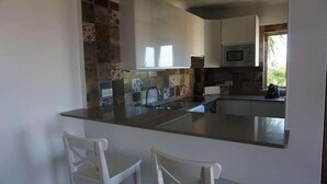 A modern kitchen with a central island, white bar stools, a microwave, and a window providing natural light. - Pontalaia Apartamentos Turisticos (Vila do Bispo)