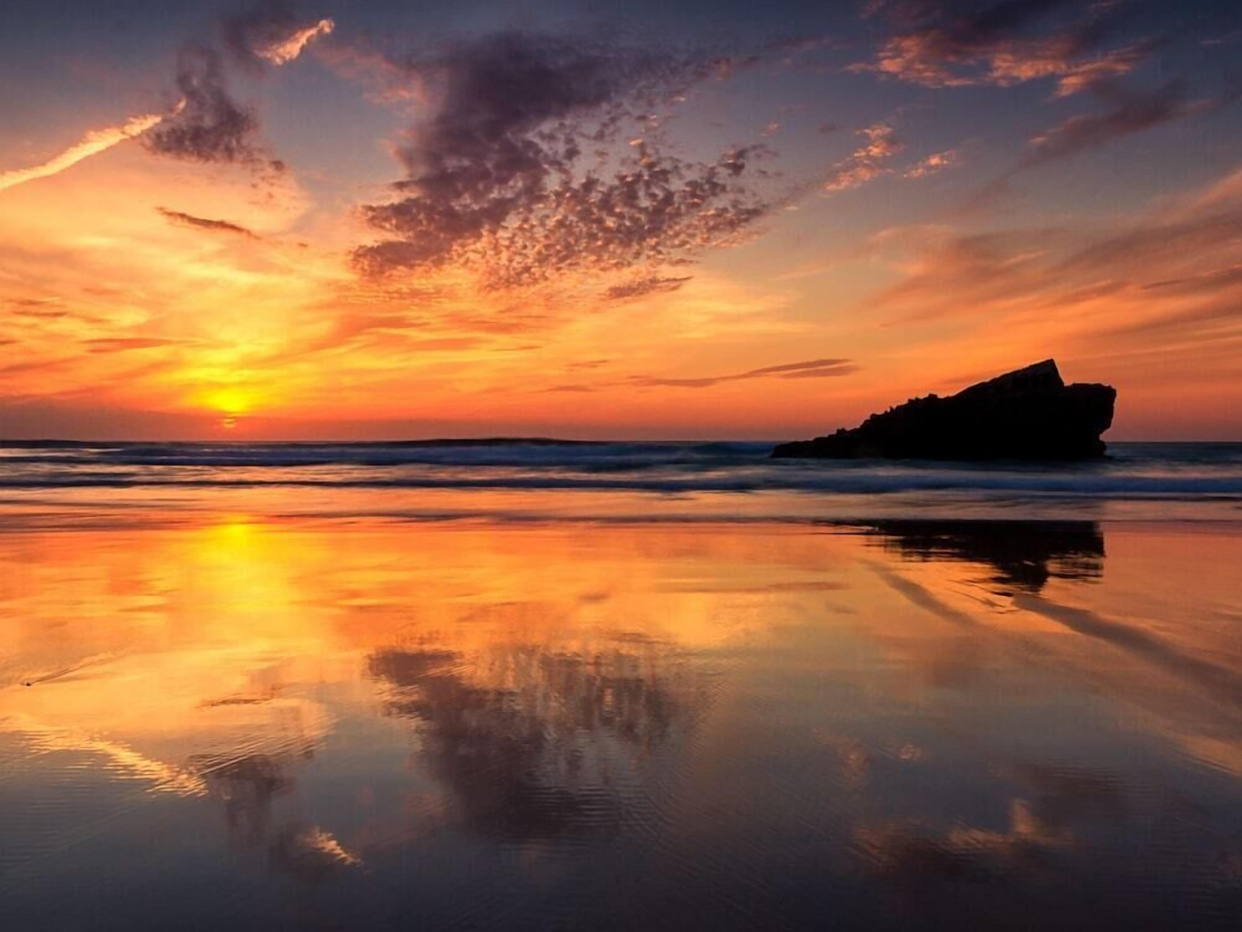 A beach scene with a rock formation and calm water reflecting the sunset.