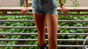 A person standing on a balcony with a metal railing, wearing a brown top, denim shorts, and cowboy boots.