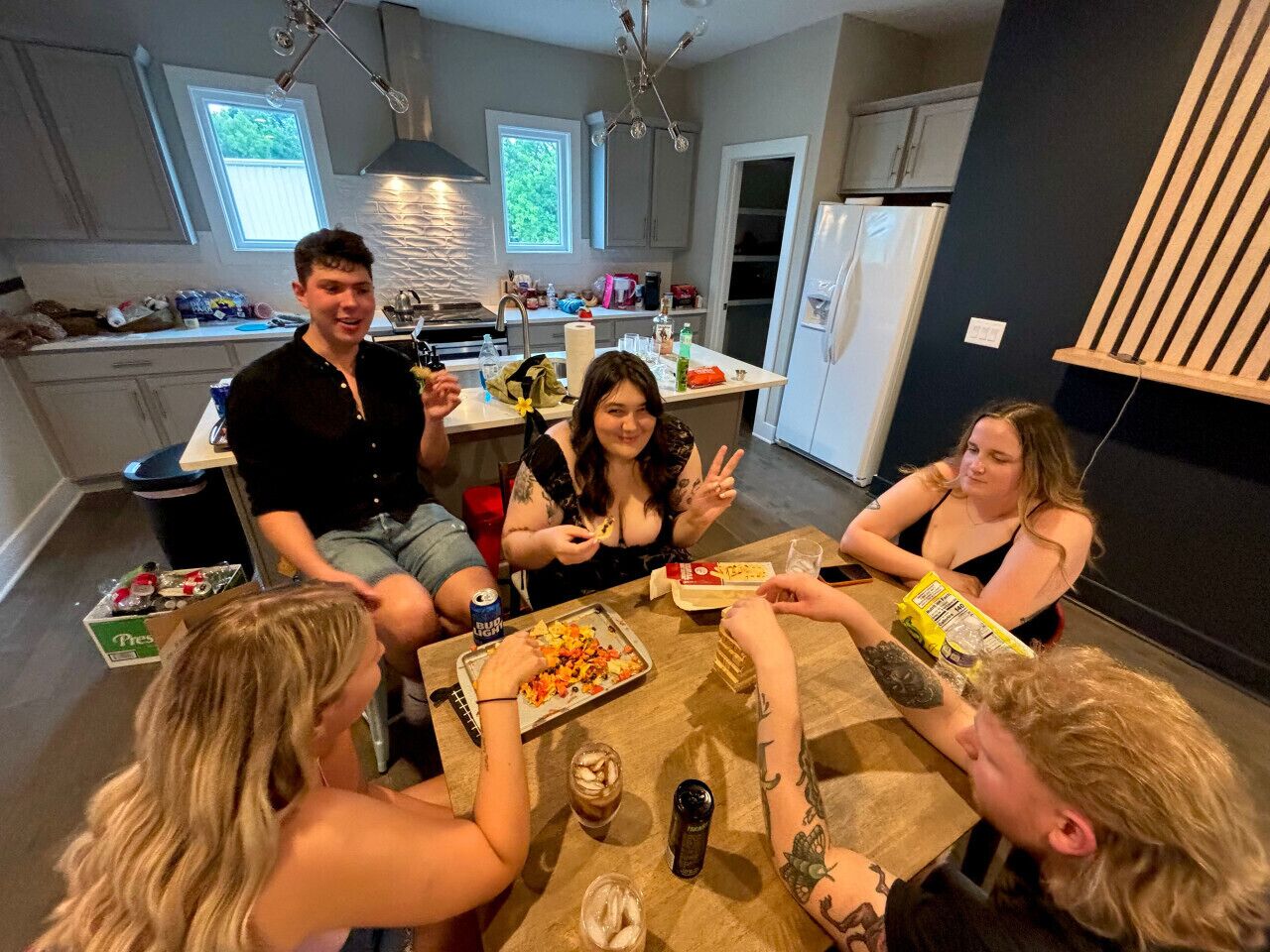 A group of people sitting around a table in a modern kitchen with a large window, a refrigerator, and a dining area.