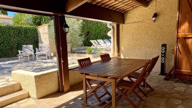 Een terras buiten met een houten tafel en stoelen, stenen vloer en uitzicht op een tuin met loungestoelen.