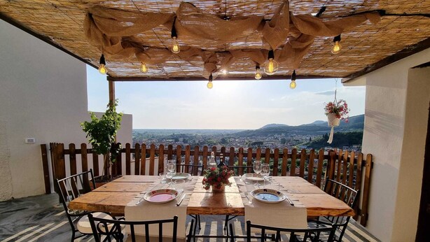 An outdoor dining area with a table set for two, surrounded by chairs, under a thatched roof with hanging lights. - House Chez Paul (Massarosa)