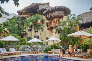 A pool area with lounge chairs and umbrellas, surrounded by tropical plants and a building with balconies. - La Villa Luz (Zihuatanejo)