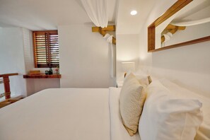 A neatly made bed with white linens and a wooden headboard, a wooden shelf with a vase and a book, and a window with wooden blinds. - La Villa Luz (Zihuatanejo)