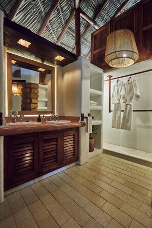 A bathroom with a large mirror, wooden vanity, two sinks, a hanging light fixture, and a towel rack with robes. - La Villa Luz (Zihuatanejo)