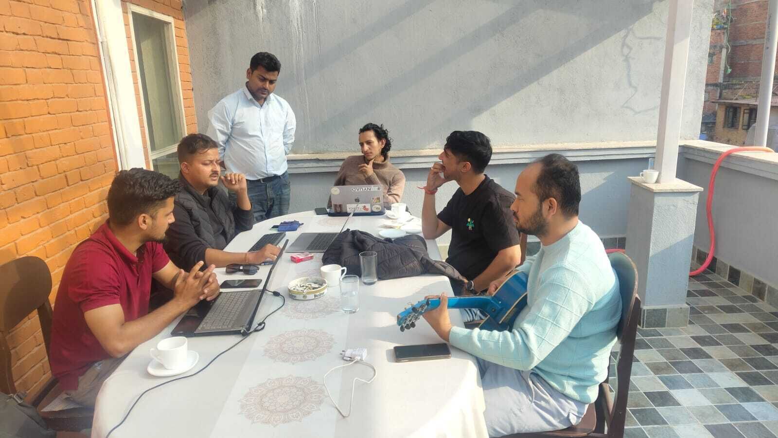 A group of people sitting around a table with laptops and a guitar, in a room with a brick wall and a window.