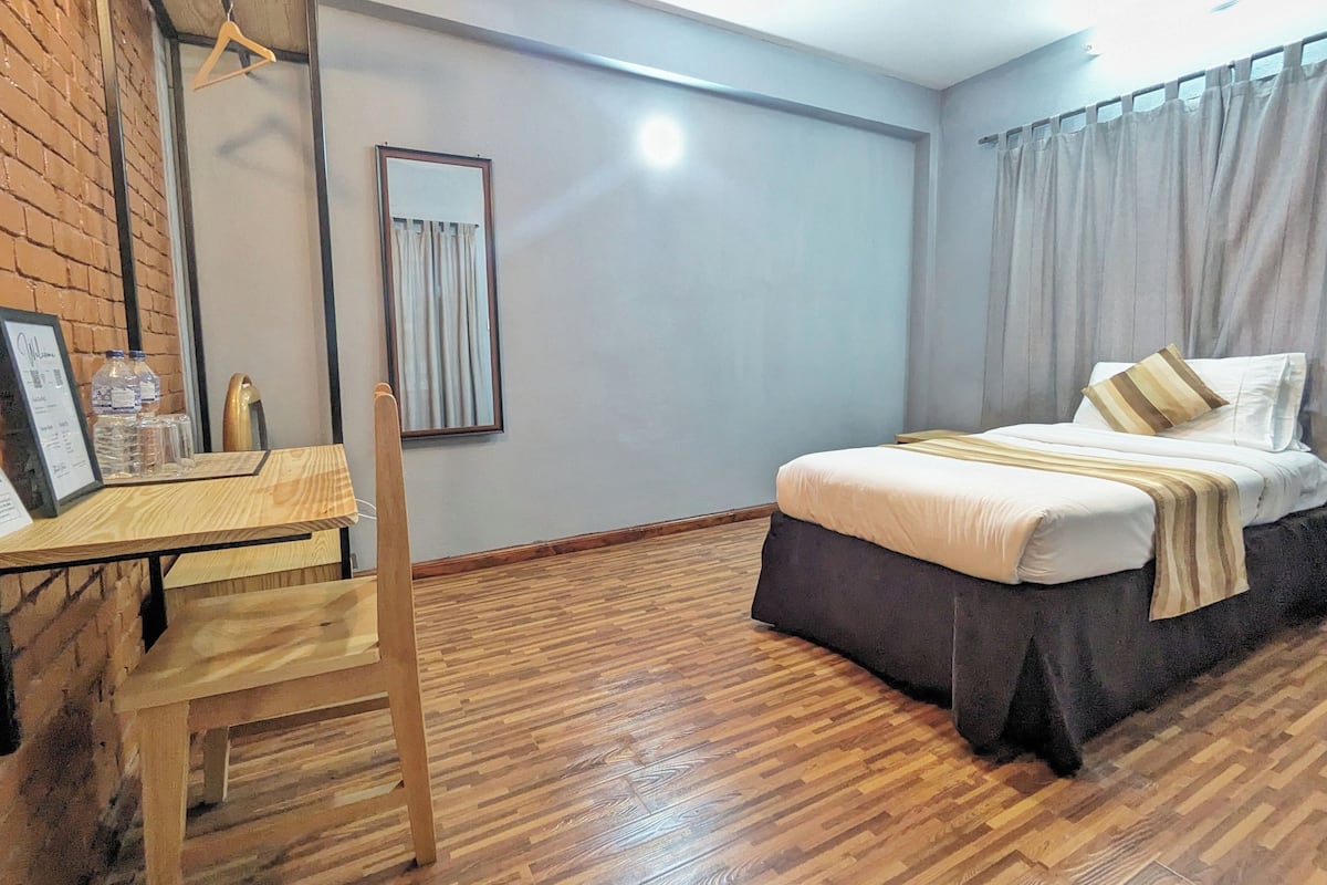 A hotel room with a wooden floor, a bed with white bedding, a desk with a chair, a framed welcome sign, and a mirror on the wall.