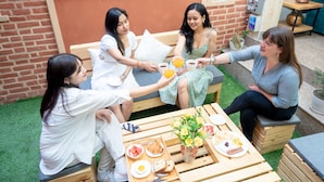 A group of people seated around a wooden table with food and drinks, in an outdoor setting with a brick wall and greenery. - Nyadha House (Lalitpur)