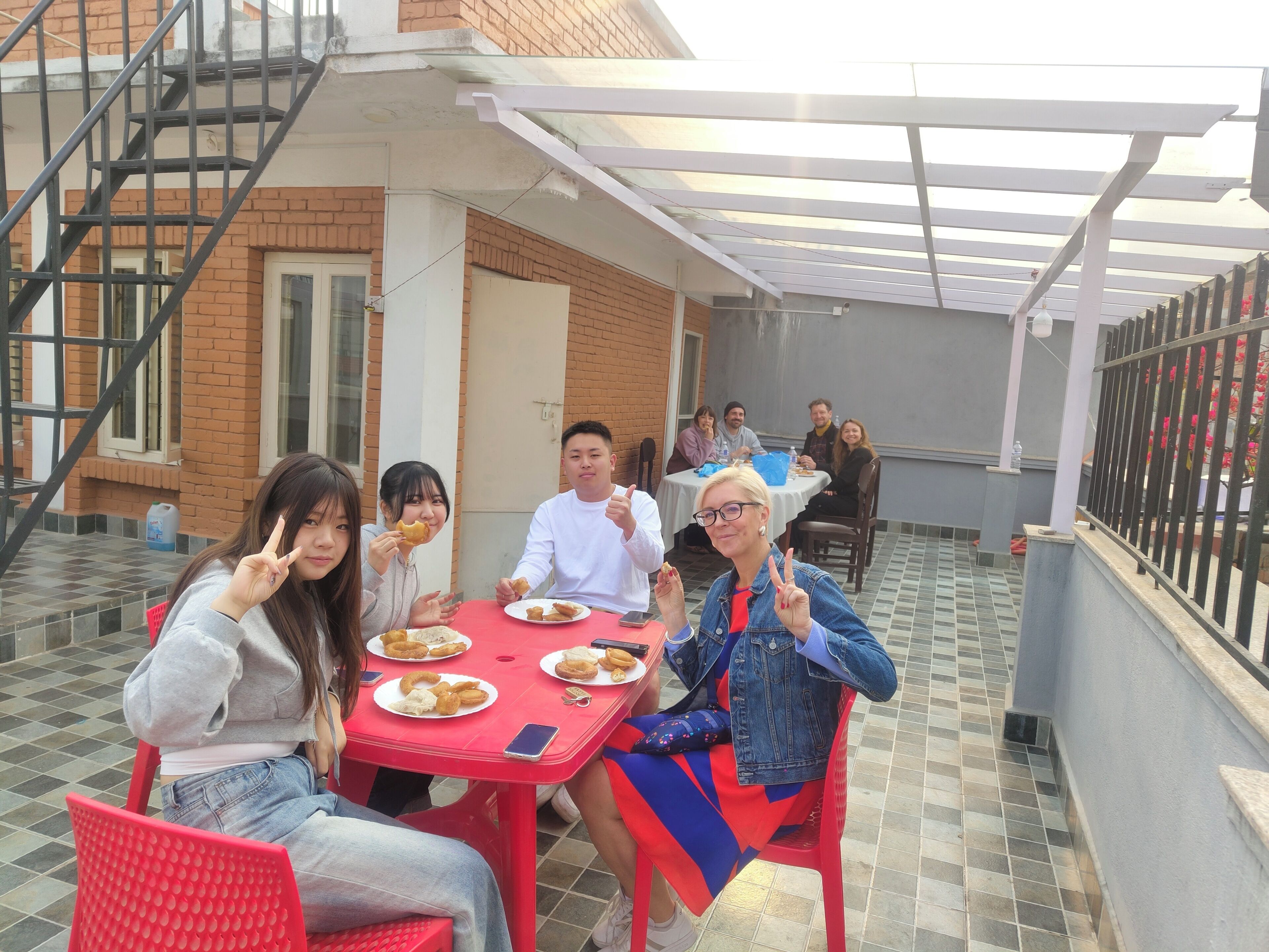 Un grupo de personas sentadas en una mesa roja al aire libre, con un edificio de ladrillo en el fondo.