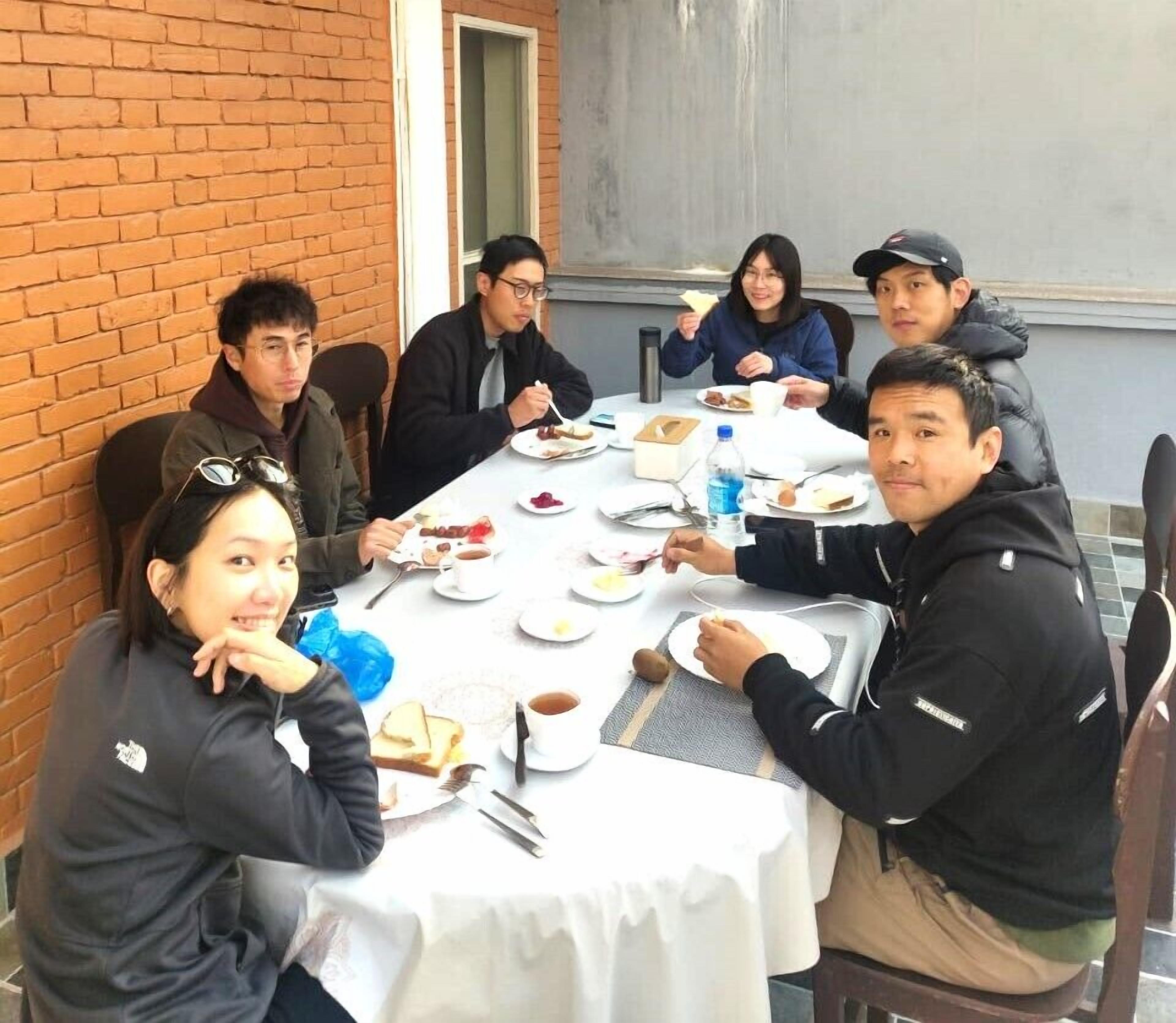 A group of people seated around a table with food and drinks, in an outdoor setting with a brick wall and a window in the background.