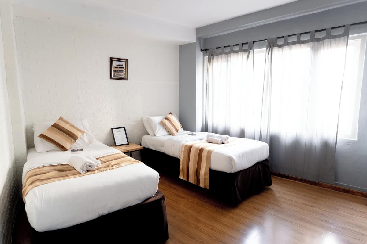 A hotel room with two beds, wooden flooring, and a window with curtains.