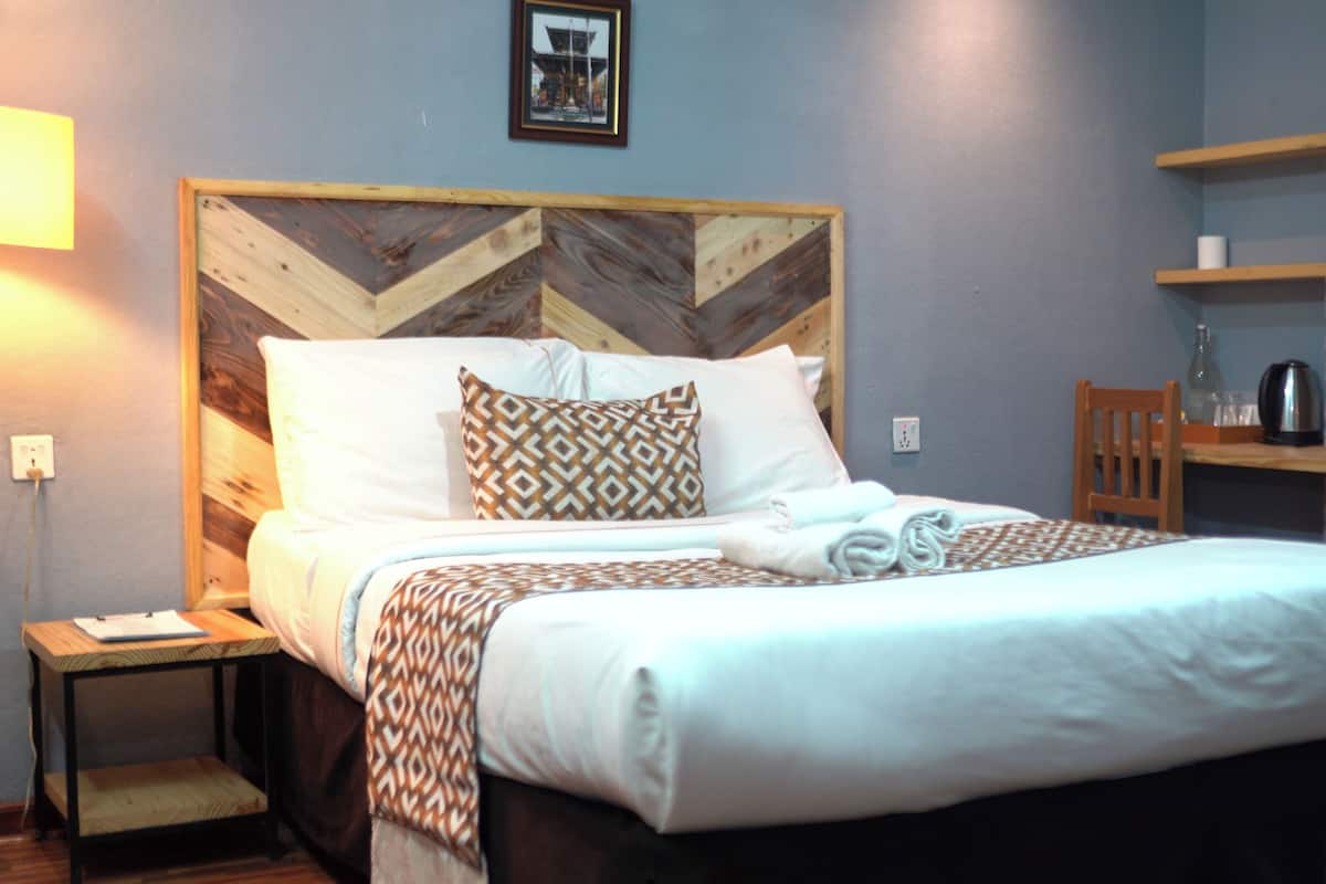 A neatly made bed with patterned bedding, a wooden headboard, a bedside table with a lamp, and a small shelf with a kettle.