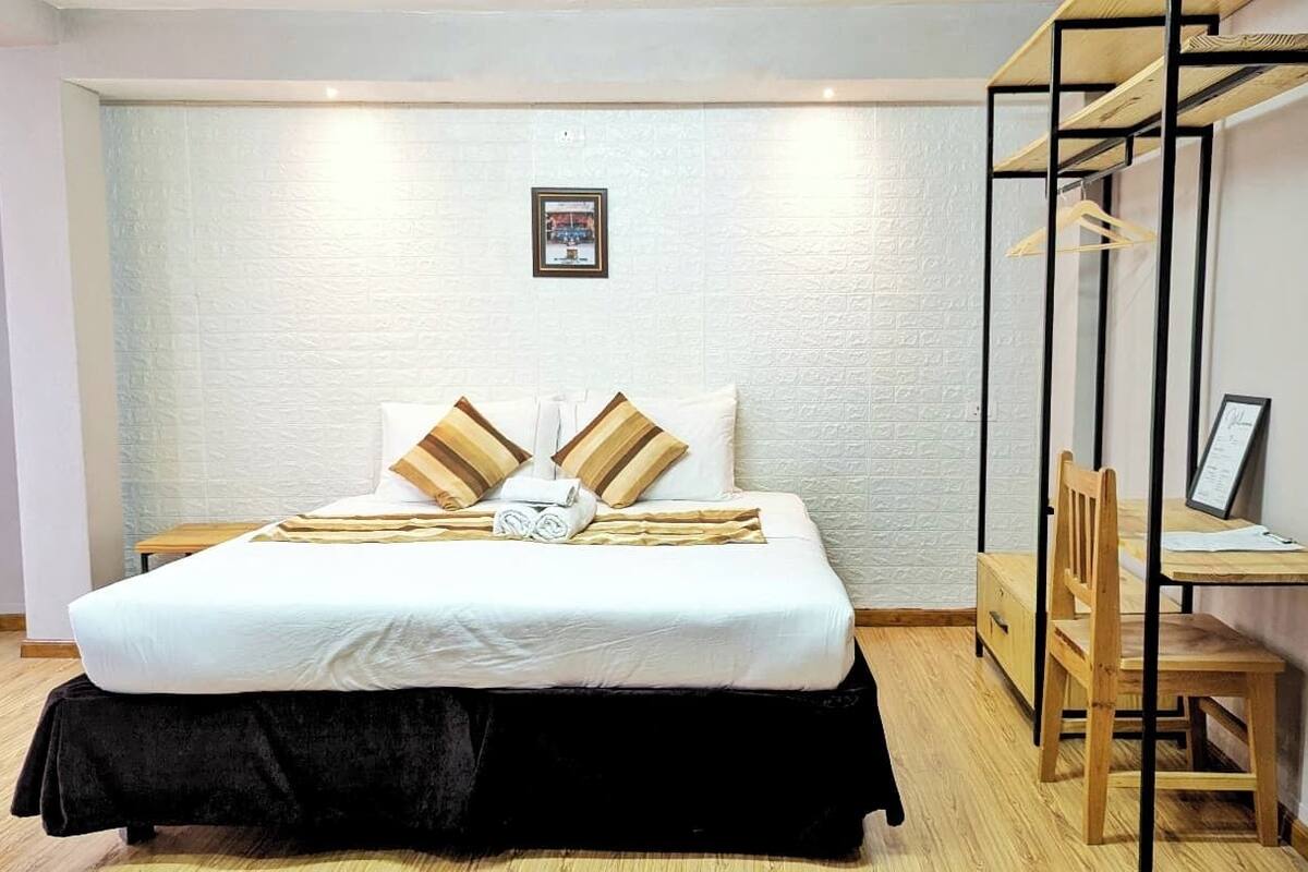A neatly made bed with a black skirt, two pillows, and a folded towel. There is a wooden chair and a desk with a computer.