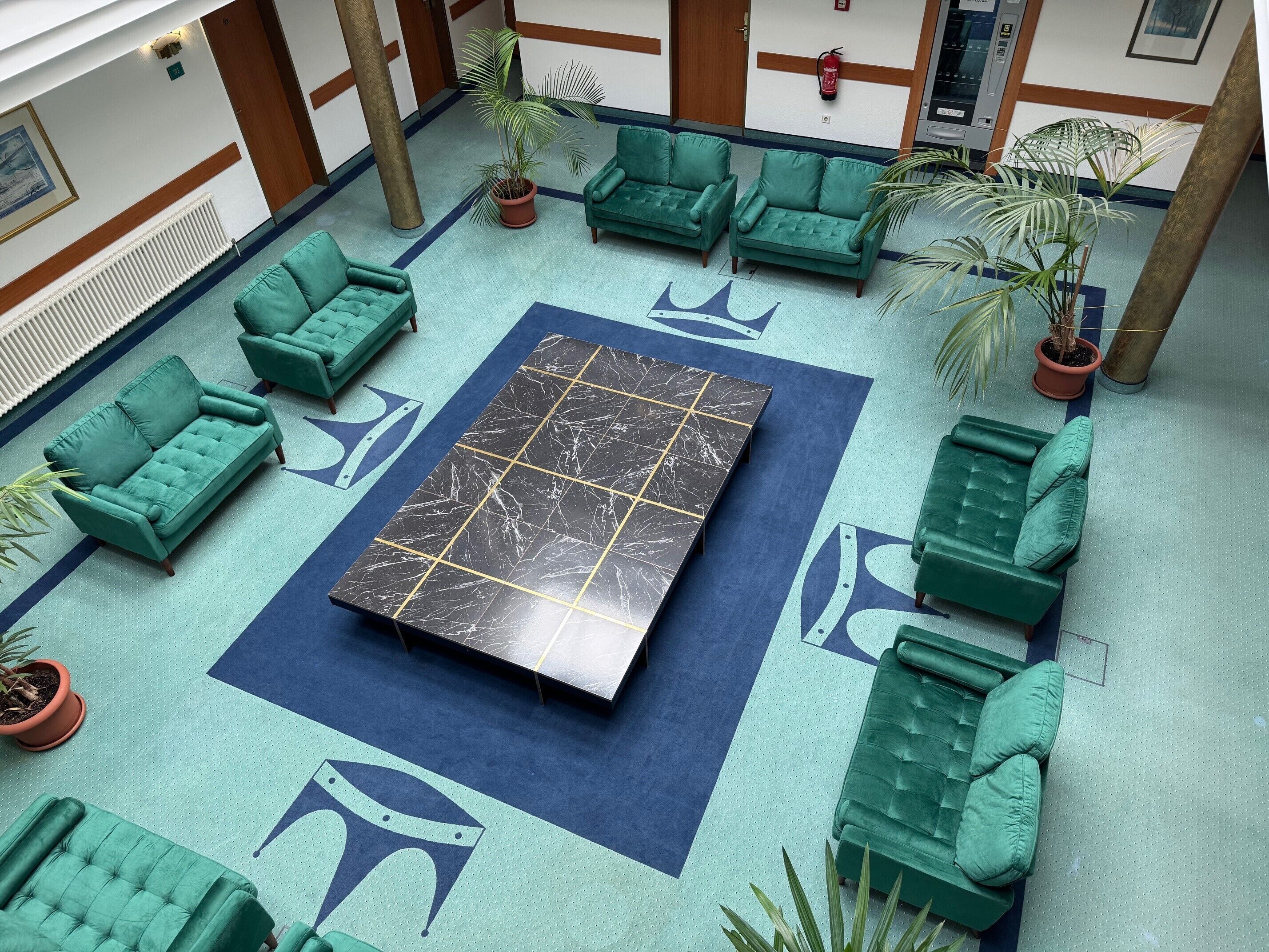 Un vestíbulo con sofás verdes, una mesa central de mármol negro y plantas en macetas.