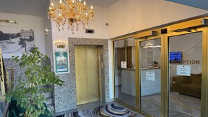 Un hall d'hôtel doté d'un lustre moderne, d'un comptoir d'accueil, d'une enseigne indiquant la réception et d'une salle de réception visible à travers des portes vitrées.