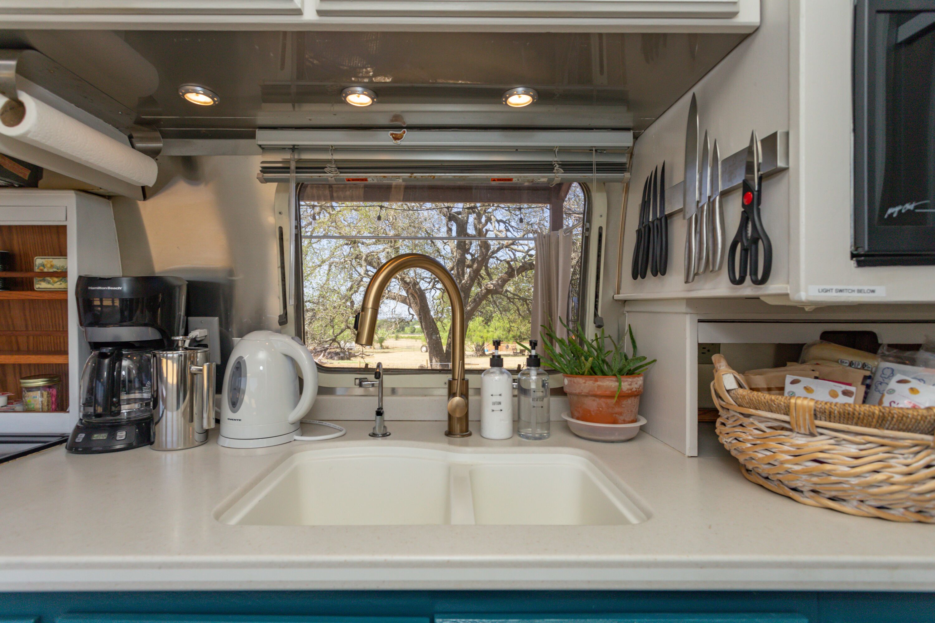 A kitchen with a sink, coffee maker, kettle, and a window with a view of trees.
