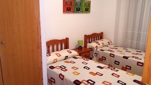 A small, cozy room with two wooden beds, each with patterned bedding. There is a wooden nightstand between the beds, and a green lamp on the nightstand. The room has a wooden door and a white wall with a framed picture above the beds. - Apartamentos Oropesa sin Piscina 3000 (Oropesa del Mar)