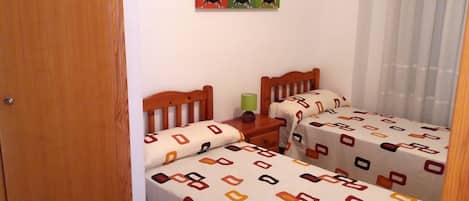 Una habitación pequeña y acogedora con dos camas de madera, cada una con ropa de cama estampada. Hay una mesita de noche de madera entre las camas, y una lámpara verde sobre la mesita. La habitación tiene una puerta de madera y una pared blanca con un cuadro enmarcado sobre las camas.