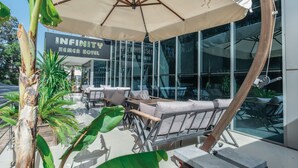 A modern hotel exterior with outdoor seating, large windows, and a sign reading "INFINITY KEMER HOTEL".