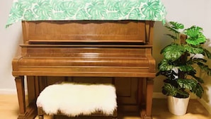 A wooden piano with a white furry ottoman and a potted plant beside it.