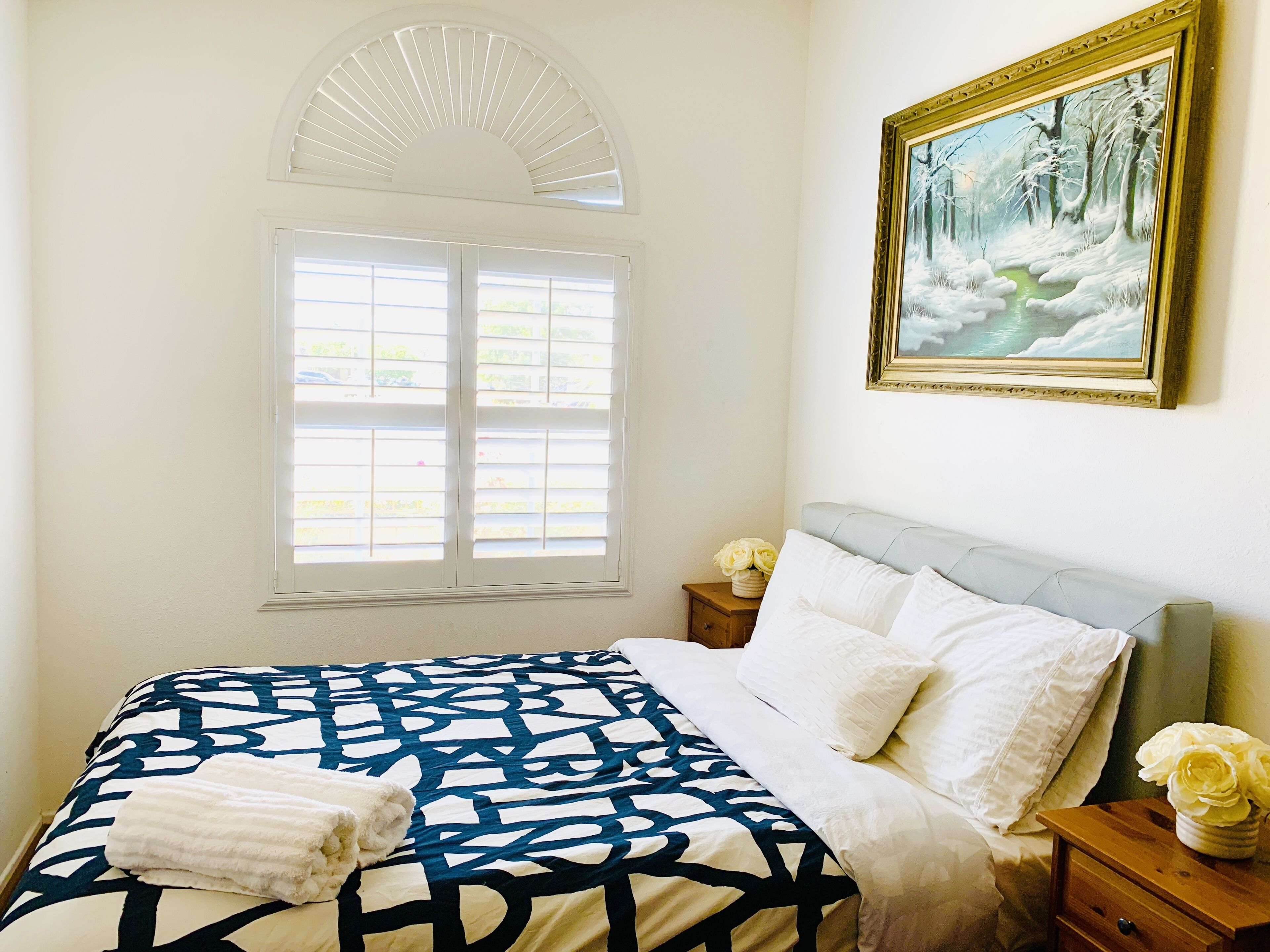A bedroom with a bed, a nightstand, a window with blinds, a painting, and a vase with flowers.