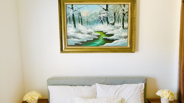 A bedroom with a bed, two nightstands, a painting of a winter scene, and a vase with flowers on one nightstand.