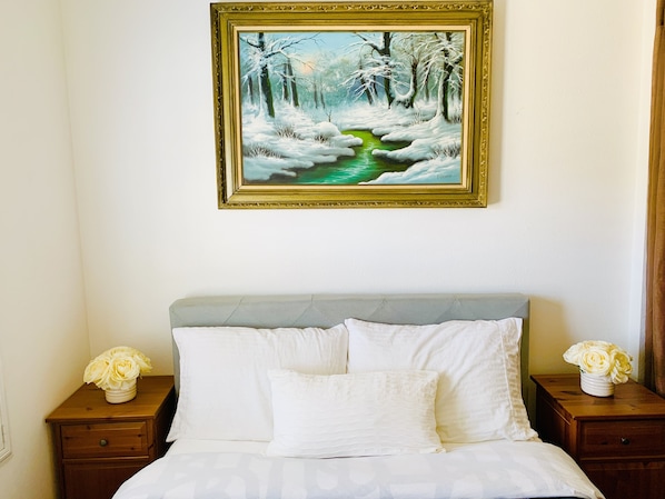 Une chambre à coucher avec un lit, deux tables de chevet, un tableau représentant un paysage hivernal et un vase rempli de fleurs posé sur l’une des tables de chevet.