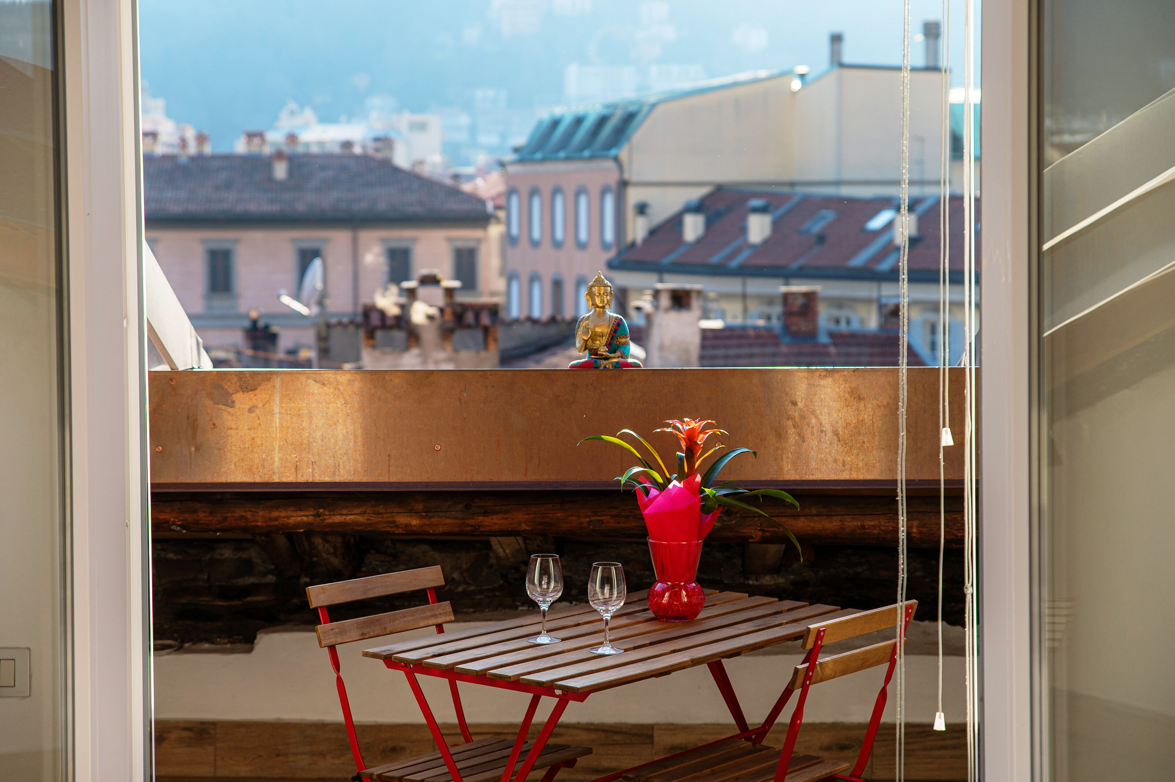 Ein Balkon mit Tisch, zwei Gläsern und einer roten Vase mit Blumen.
