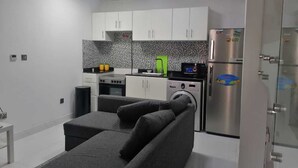 A compact kitchen with white cabinets, a stainless steel refrigerator, a washing machine, and a grey sofa.