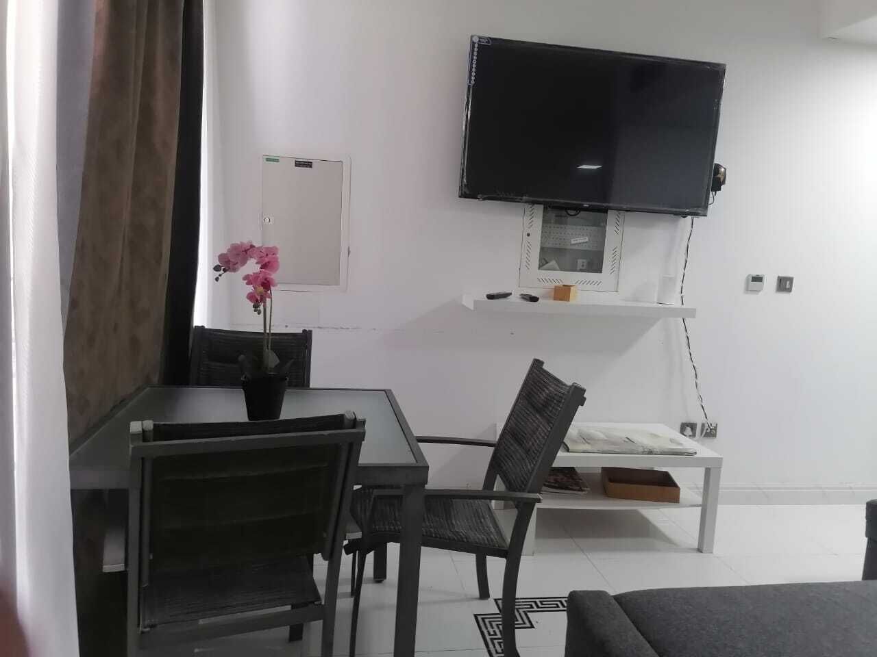 Una sala de estar moderna con una televisión de pantalla plana montada en la pared, una mesa de comedor con sillas, un sofá y una pequeña planta en maceta.