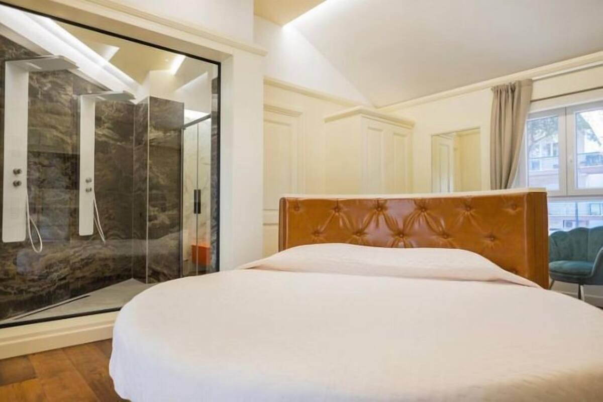 A bedroom with a large bed, a glass shower enclosure, and a view of the outside.