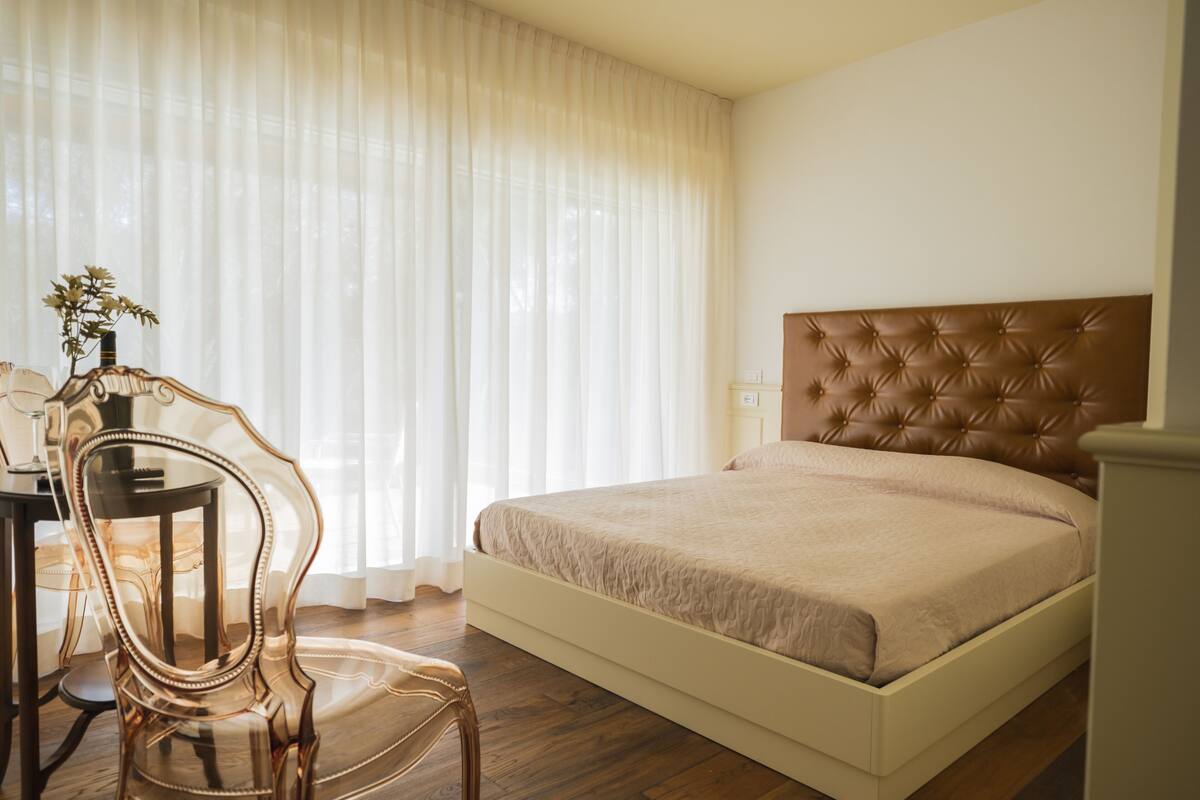 A bedroom with a bed, a chair, a table, and a window with sheer curtains.