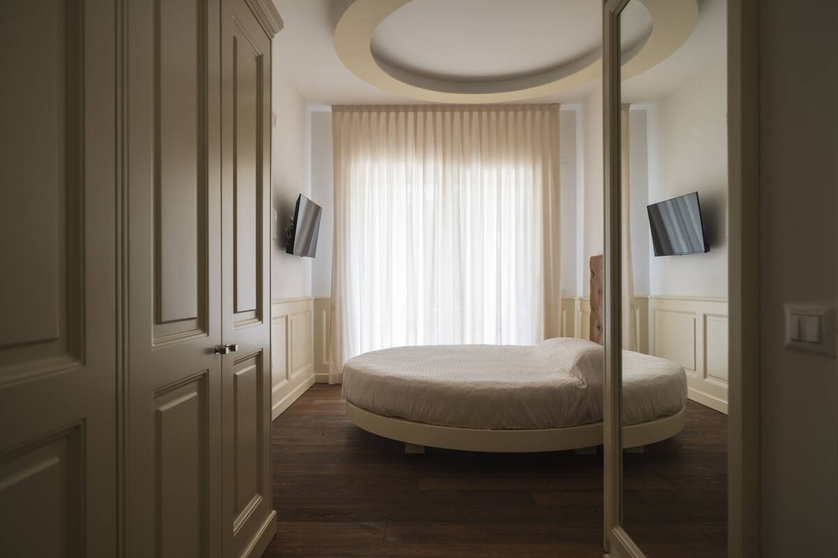 A bedroom with a round bed, a large window with curtains, and two wall-mounted TVs.