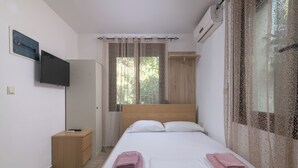 A bedroom with a bed, a TV mounted on the wall, a wooden wardrobe, and a window with curtains. - Parko Studios - Studio 1 (Rodos)