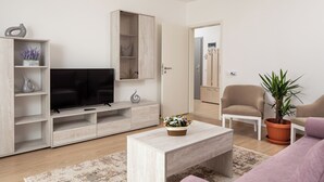 A modern living room with a flat-screen TV, wooden shelving, a coffee table, a potted plant, and a beige armchair. - Brasov Holiday Apartments-purple (Brasov)