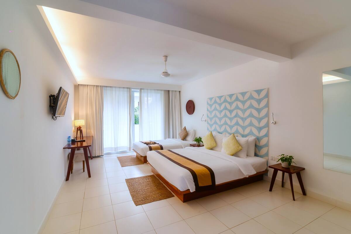 A hotel room with two beds, a desk with a lamp, a wall clock, and a large window with curtains.