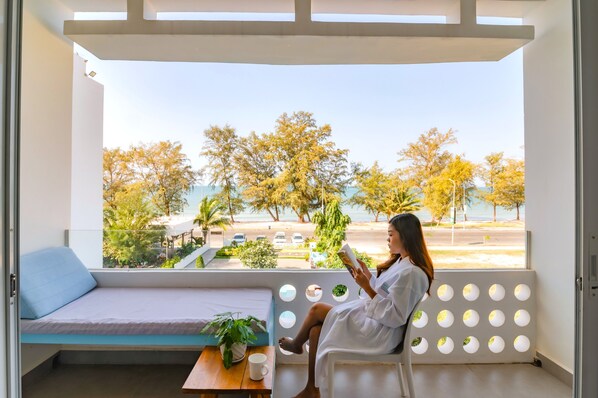 A balcony with a view of trees and a road, a white sofa, a table with a plant, and a person reading a book. - Naiya Sea Resort (Sihanoukville)