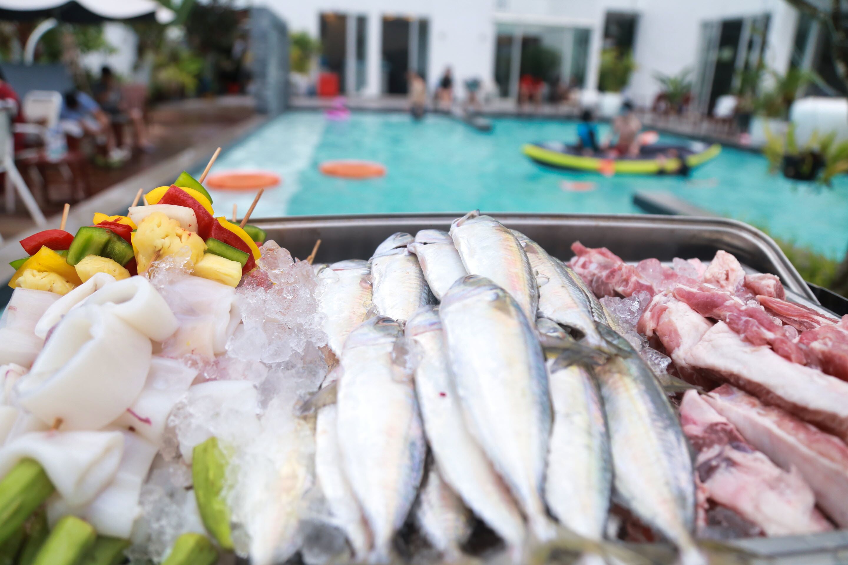 Exhibición de mariscos con diversos peces y verduras sobre hielo, una piscina con gente y un bote al fondo.