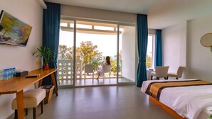 Ein Hotelzimmer mit Bett, Schreibtisch, Sessel, Fernseher und einem Balkon mit Blick auf Bäume.