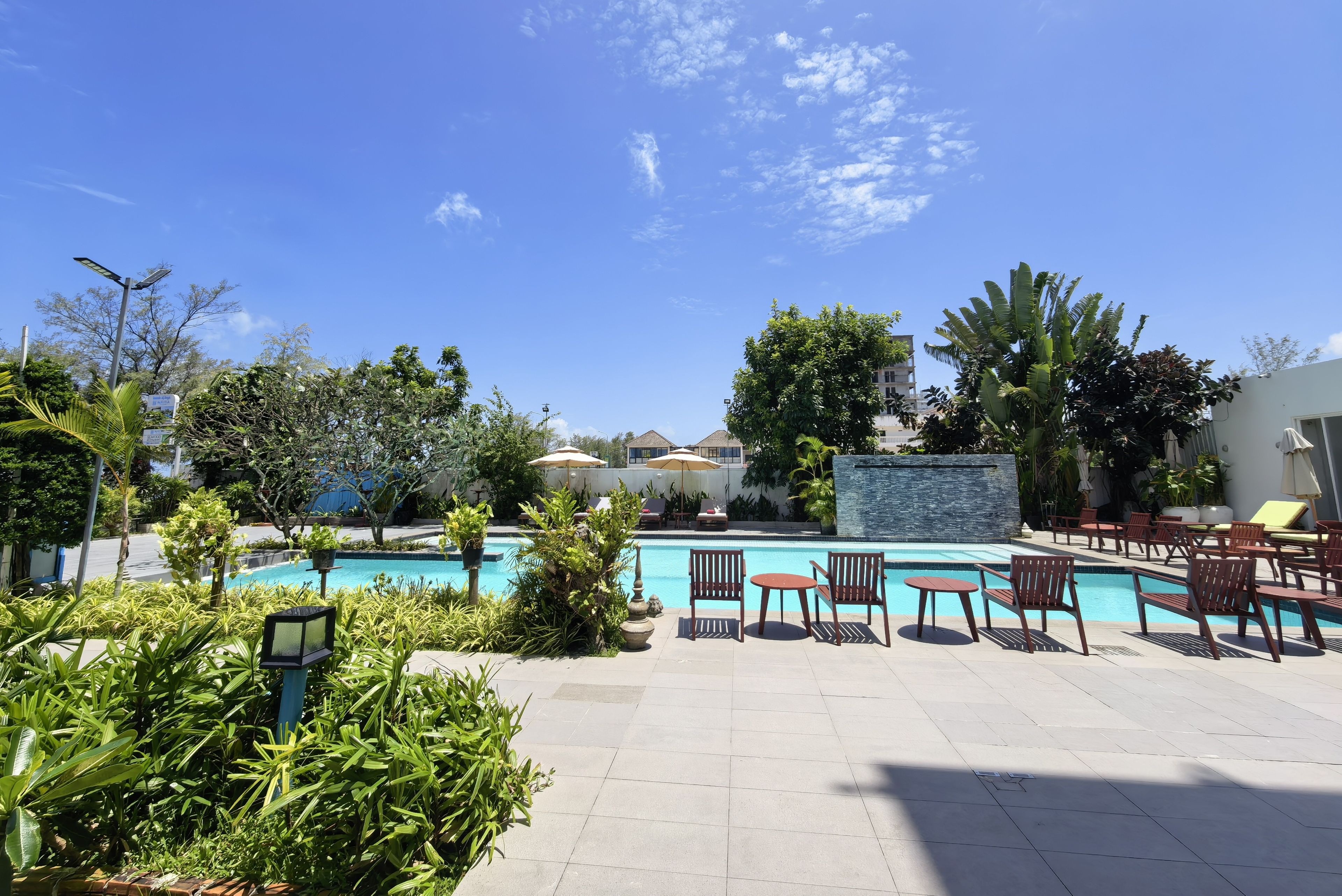 A pool area with lounge chairs and umbrellas, surrounded by greenery and a clear sky.