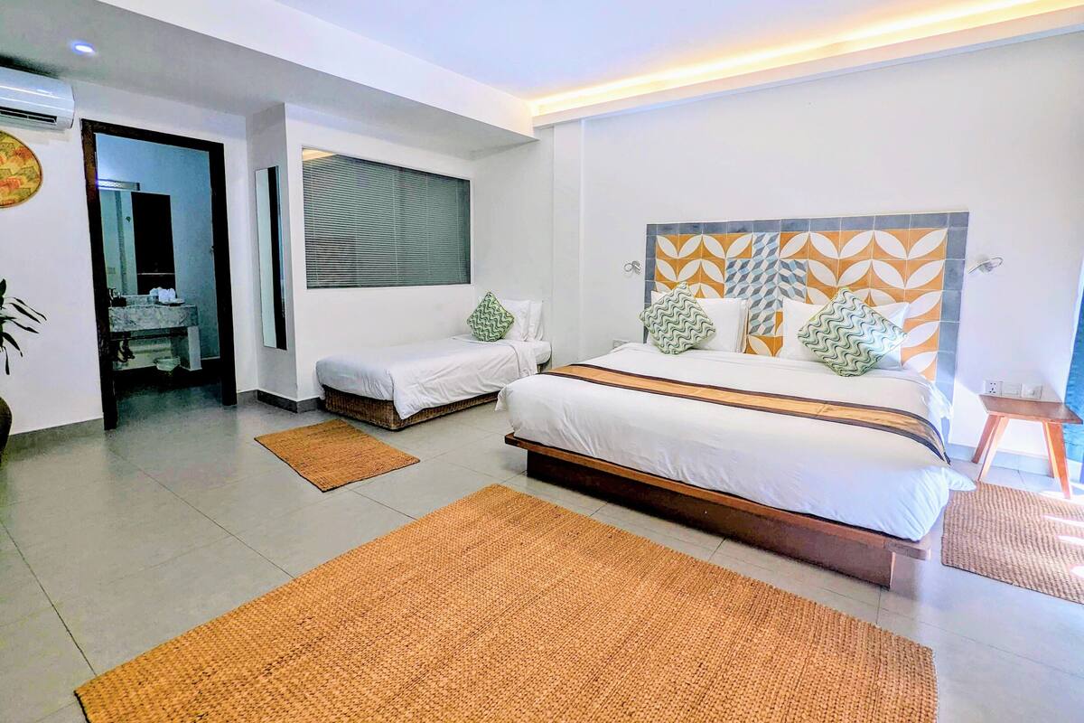A modern hotel room with a large bed, two smaller beds, a bathroom with a mirror, and a view of the outdoors.