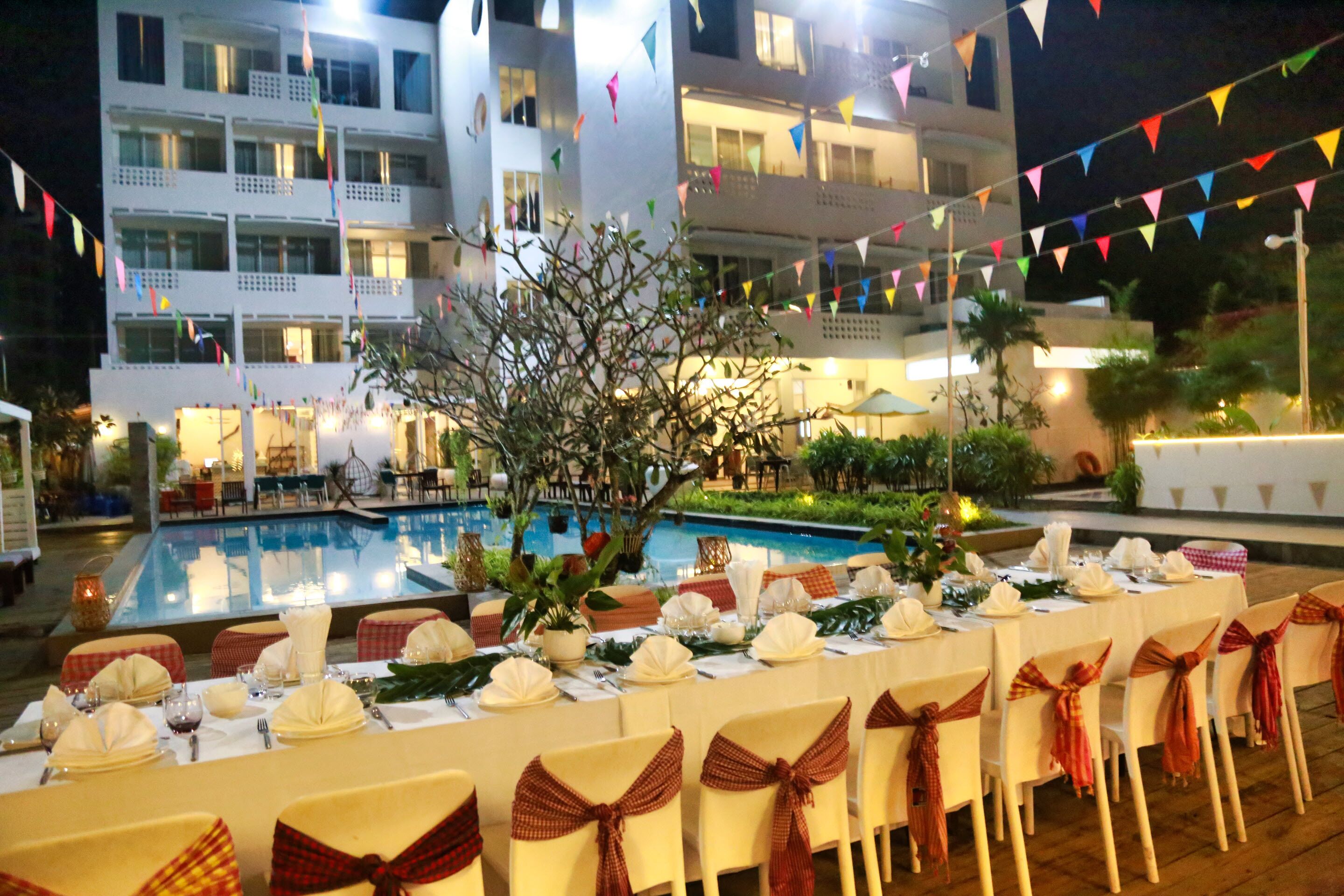 Una mesa de banquete dispuesta para un evento formal, con sillas decoradas con fajas a cuadros rojos y blancos, una piscina al fondo y un edificio con balcones y banderas coloridas.