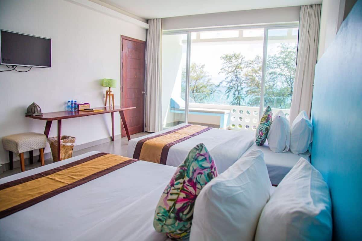 A hotel room with two beds, a desk, a television, and a balcony with a view of trees.