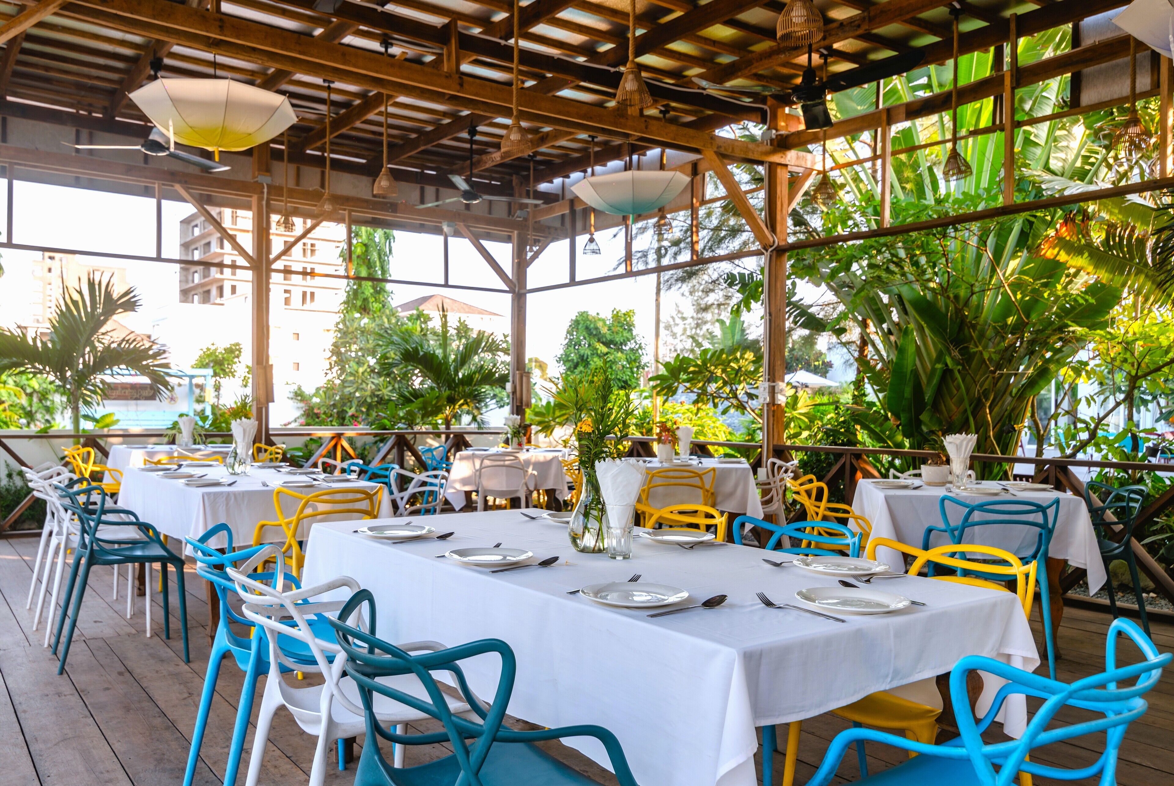 Restaurante ao ar livre com mesas e cadeiras, coberto por um telhado de madeira, rodeado por vegetação.
