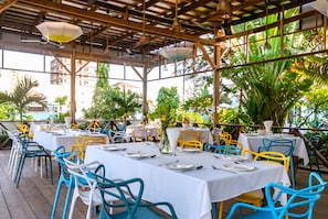An outdoor restaurant with tables and chairs, covered by a wooden roof, surrounded by greenery.