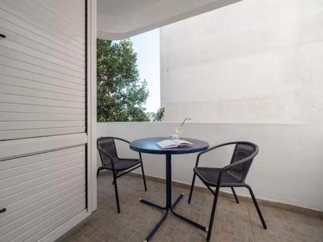 Un balcón con una mesa redonda y dos sillas, una pared blanca y un árbol al fondo.
