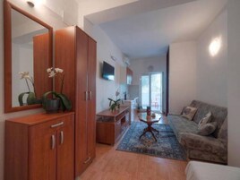 Ein modernes Wohnzimmer mit einer Couch, einem Couchtisch und einem Schrank. Ein Fernseher ist an der Wand montiert und ein Fenster lässt natürliches Licht herein.