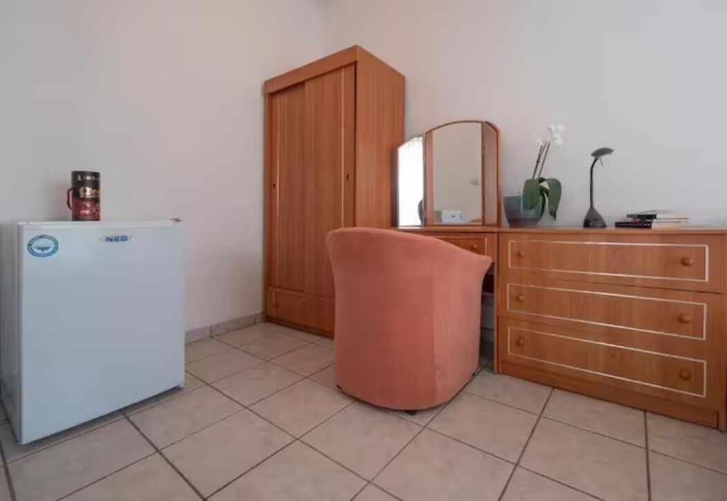 A room with a white refrigerator, a wooden wardrobe, a pink chair, a wooden dresser, and a mirror.